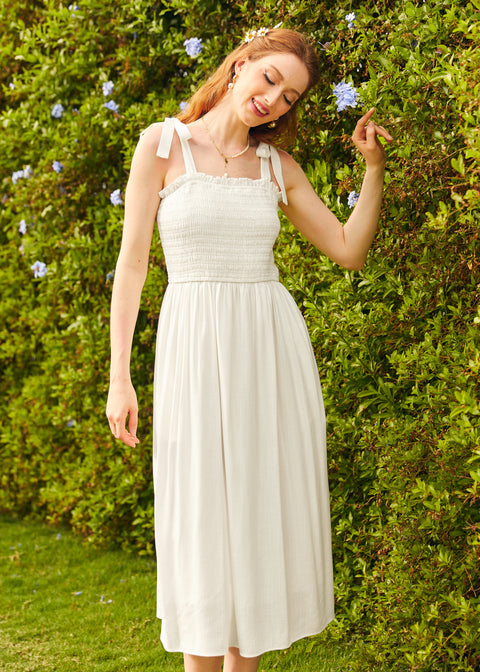 A woman standing in front of a hedge wearing a white midi dress with tie-bow shoulder straps and a square neckline.