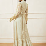 Woman wearing a long, light-colored dress with lace sleeves against a white wall.