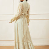 Woman wearing a long, light-colored dress with lace sleeves against a white wall.