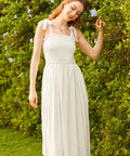 A woman standing in front of a hedge wearing a white midi dress with tie-bow shoulder straps and a square neckline.