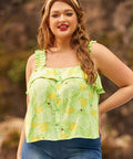 A woman wearing a light green, lemon-patterned blouse with ruffled spaghetti straps and a button-down front, paired with blue jeans.