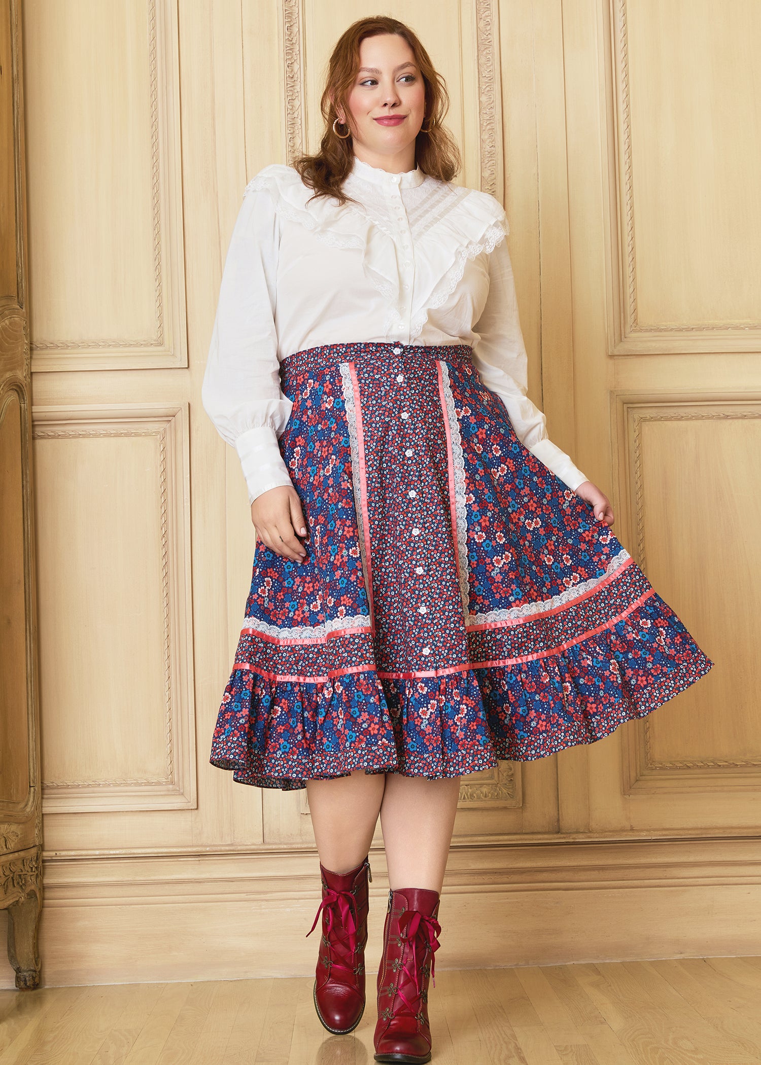 A woman stands against a wooden background wearing a floral A-line midi skirt with lace and satin ribbon trim, flower buttons, and a white blouse with ruffled details.
