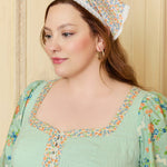 Woman wearing a floral dress and headscarf against a beige wall.