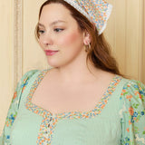Woman wearing a floral dress and headscarf against a beige wall.
