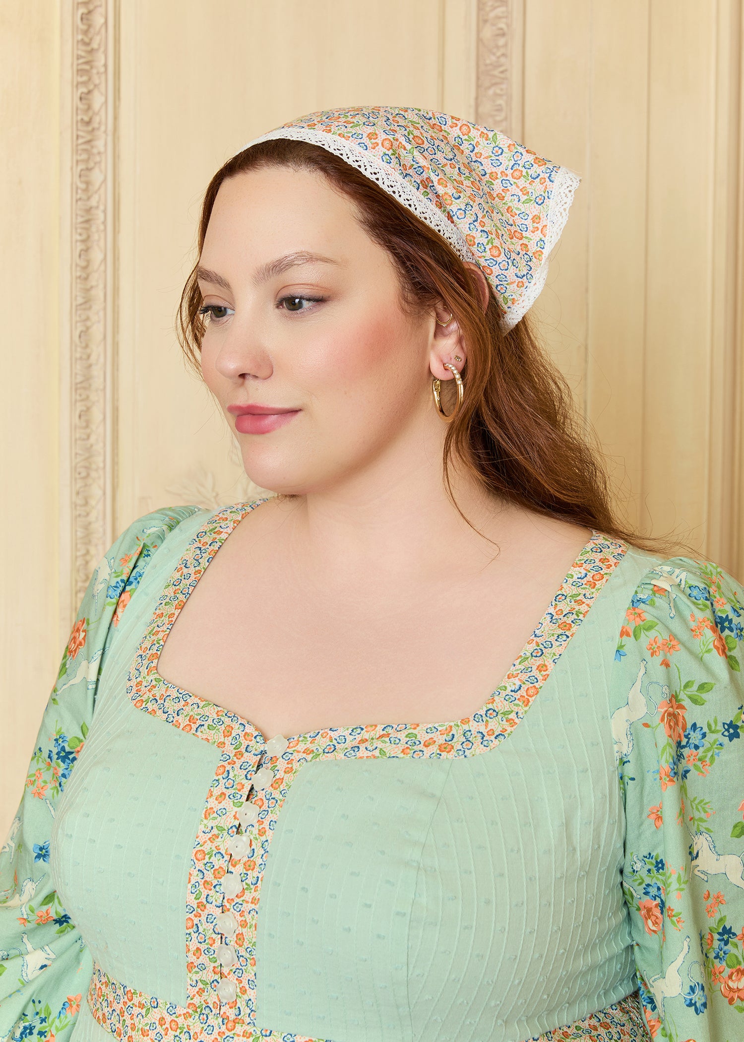 Woman wearing a floral dress and headscarf against a beige wall.