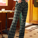 A person standing in a cafe wearing high-waisted 70's inspired green plaid wide-leg trousers with pockets.