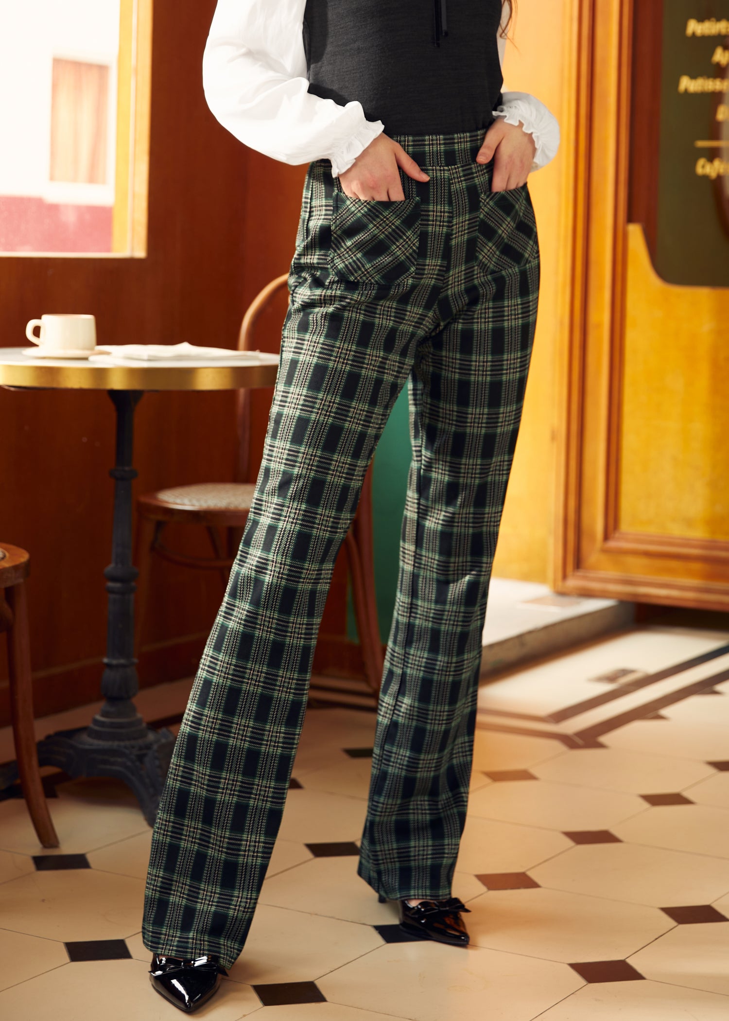 A person standing in a cafe wearing high-waisted 70's inspired green plaid wide-leg trousers with pockets.