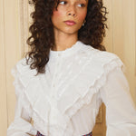 A woman wearing a white Edwardian-style ruffle blouse with a high neckline and long cuffed sleeves, paired with a multicolored skirt.