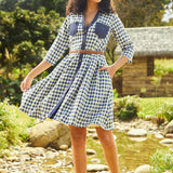 A woman standing outdoors in a casual chic setting, wearing a blue and white checkered shirtdress with long sleeves, a round neckline, and a belt.