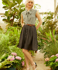A woman standing in a garden setting, wearing a vintage checkered bow blouse with cap sleeves and a high-waisted skirt.