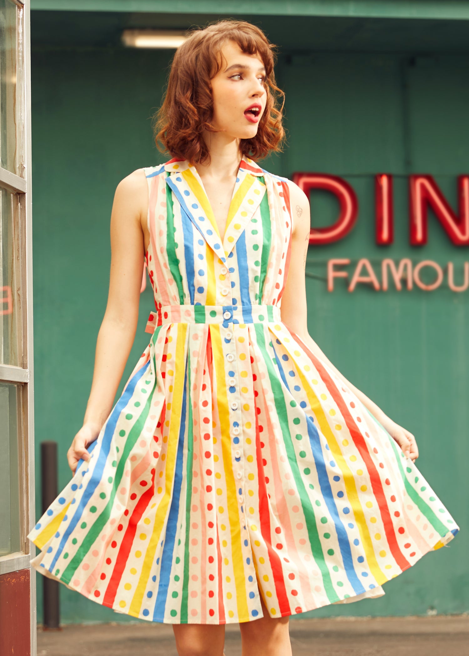 Woman wearing sleeveless rainbow striped polka dot shirt dress with button-front bodice and fit-and-flare skirt.