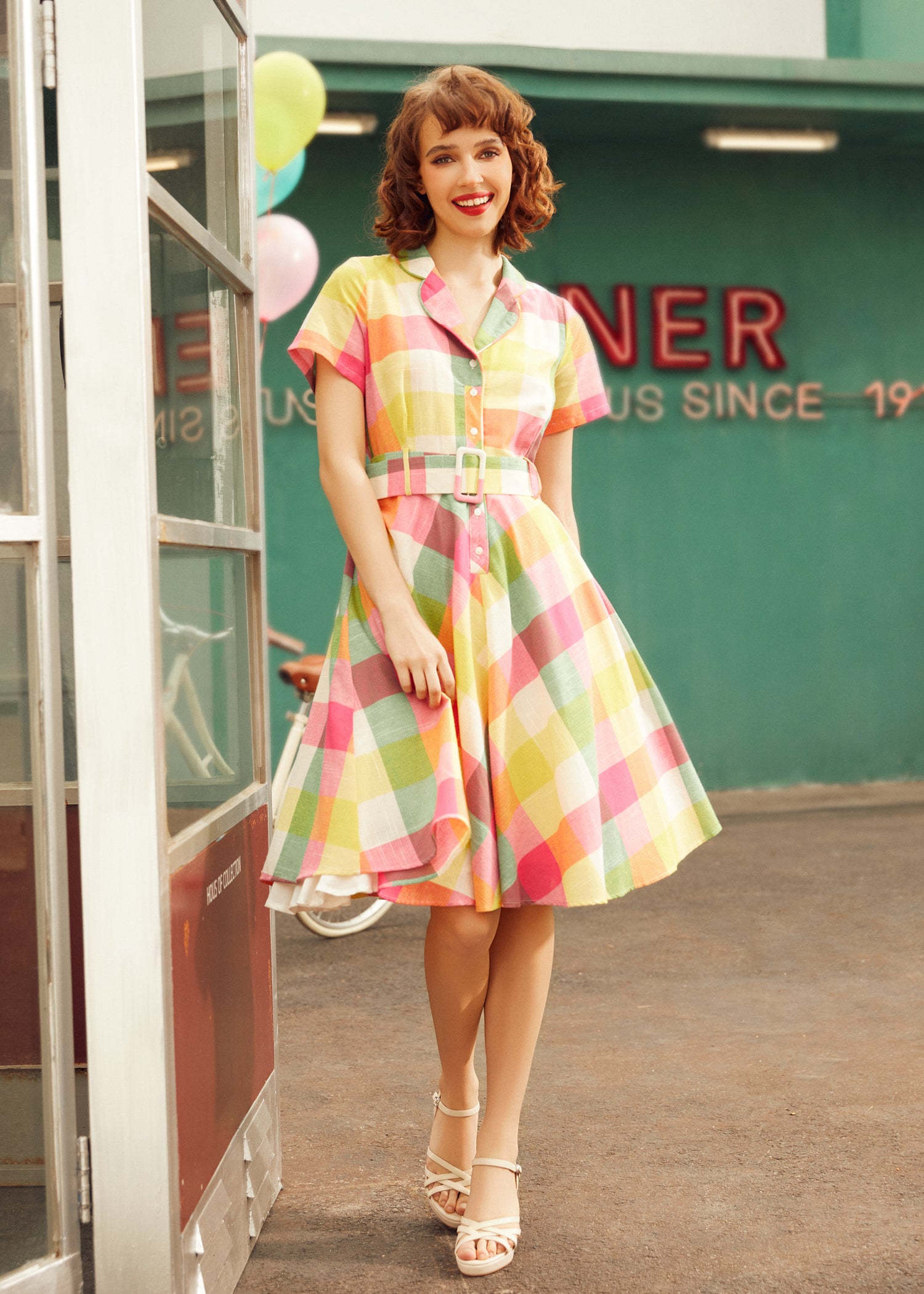 Woman wearing a colorful pink, yellow, green, and orange plaid dress with a button-front bodice.