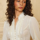 Woman wearing a white blouse with intricate details against a beige wall.