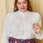 A woman wearing a white high-necked, long-sleeved blouse with ruffled detailing on the bodice and trims on the sleeves. She is also wearing a multicolored skirt with a floral pattern.