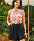 Woman wearing a floral sleeveless top and black shorts standing outdoors with greenery in the background.