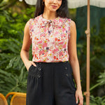 Woman wearing a floral sleeveless top and black shorts standing outdoors with greenery in the background.