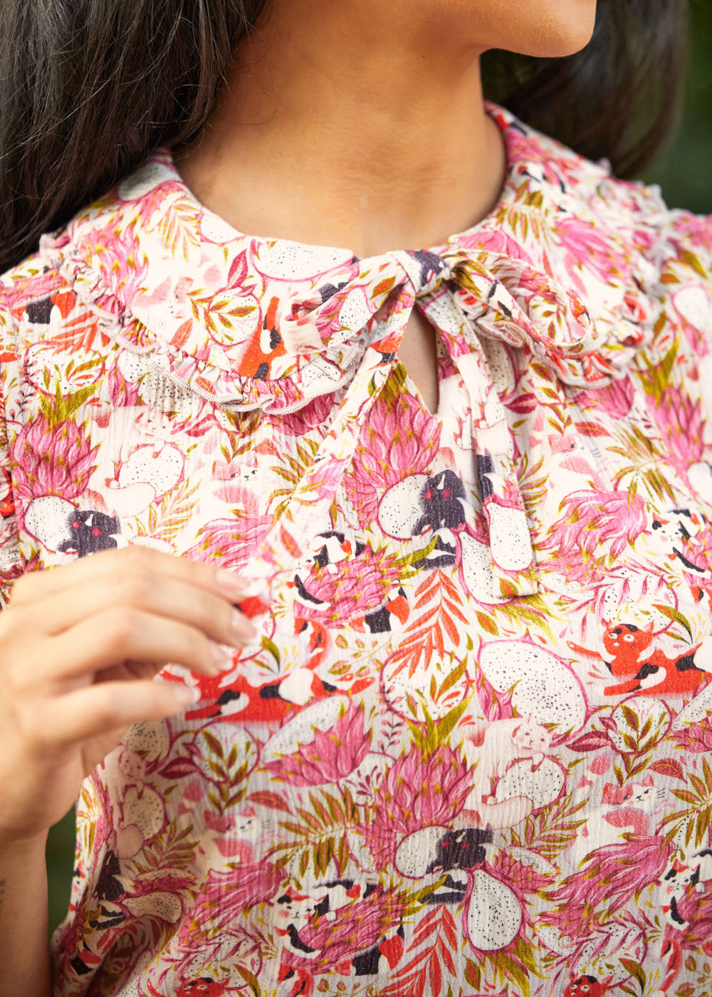 Close-up of a person wearing a floral patterned blouse with dragonfruit and hidden cats, a peter pan collar with ruffle trim, and a bow.