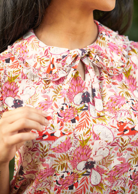 Close-up of a person wearing a floral patterned blouse with dragonfruit and hidden cats, a peter pan collar with ruffle trim, and a bow.