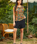Woman wearing a colorful peach fruit print top and black skirt standing outdoors with greenery and a chair in the background.