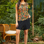 Woman wearing a colorful peach fruit print top and black skirt standing outdoors with greenery and a chair in the background.