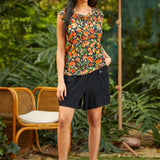 Woman wearing a colorful peach fruit print top and black skirt standing outdoors with greenery and a chair in the background.
