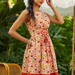 Woman wearing a patterned dress with a red bottom section, standing outdoors with greenery in the background.