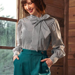 Woman wearing a striped blouse with a large bow and teal pants indoors.