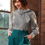 Woman wearing a striped blouse with a large bow and teal pants indoors.