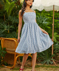 Woman in a light blue floral dress standing outdoors with greenery and a white umbrella in the background.