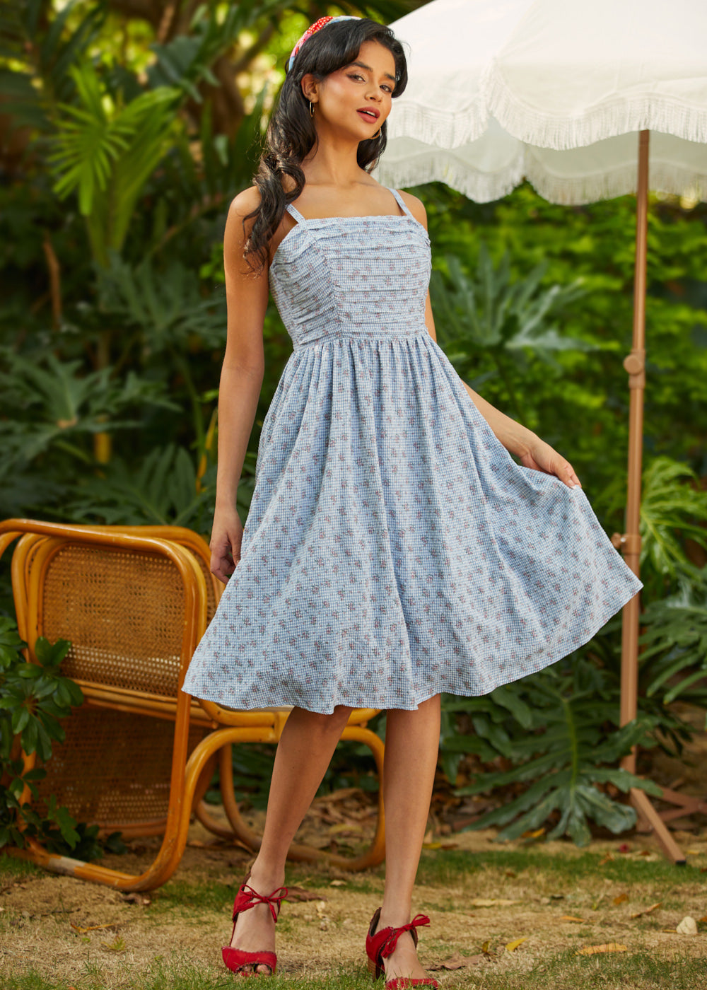 Woman in a light blue floral dress standing outdoors with greenery and a white umbrella in the background.