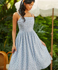 Woman in a blue patterned dress standing outdoors with greenery and an umbrella in the background