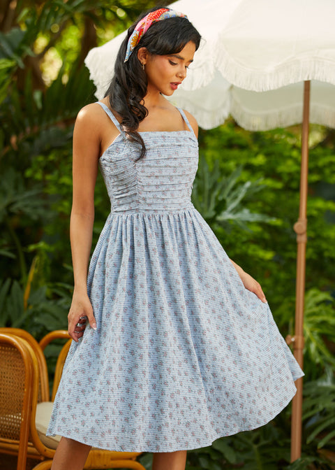 Woman in a blue patterned dress standing outdoors with greenery and an umbrella in the background