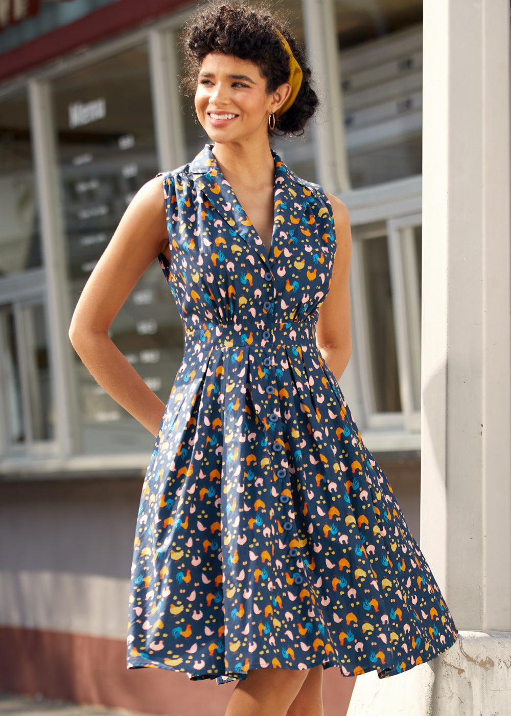 Woman wearing sleeveless chicken print dress with button-front bodice, notched collar, and gathered skirt.