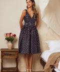 Woman in a vintage inspired polka dot dress standing in a bedroom with a vase of flowers on a table.