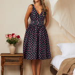 Woman in a vintage inspired polka dot dress standing in a bedroom with a vase of flowers on a table.
