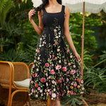 Woman wearing a black floral dress standing outdoors with greenery and a chair in the background.