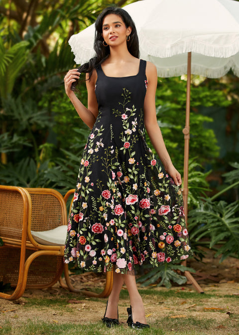 Woman wearing a black floral dress standing outdoors with greenery and a chair in the background.