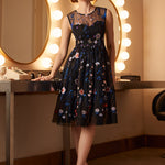 Woman wearing a black embroidered floral fit-and-flare dress with layered tulle skirt in a dressing room setting.