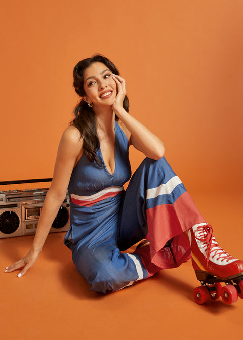 Woman seated in a blue halter jumpsuit featuring contrast stripe details and flowing legs.