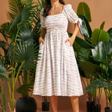 Woman wearing a white smocked square neck midi dress with colorful textured stripes, puff sleeves, and pockets.