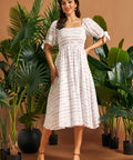 Full-length view of a woman wearing a white square neck midi dress with smocking, tie sleeves, and colorful stitched stripes.