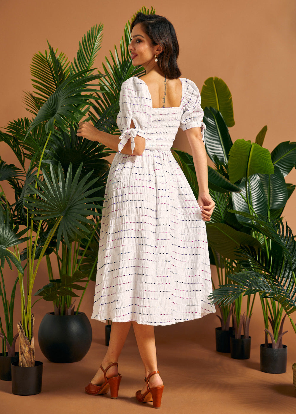 Back view of a woman wearing a white smocked midi dress with a square back neckline and tie sleeve details.