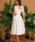 Back view of a woman wearing a white smocked midi dress with a square back neckline and tie sleeve details.
