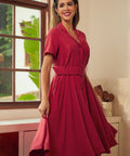A woman modeling a short-sleeve, collared, belted midi dress in a rich wine red color, standing in a brightly lit room with a window and kitchen cabinets in the background.