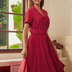 A woman modeling a short-sleeve, collared, belted midi dress in a rich wine red color, standing in a brightly lit room with a window and kitchen cabinets in the background.