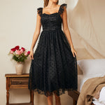 Woman in a black lace dress standing in a bedroom with a wooden side table and flowers.