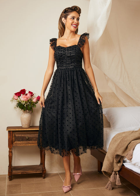 Woman in a black lace dress standing in a bedroom with a wooden side table and flowers.