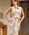 A woman modeling a sleeveless collared dress with a colorful geometric pattern in shades of yellow, blue, red, and green, featuring a button-up front and a matching tie belt at the waist.