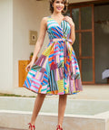 A woman modeling a sleeveless, knee-length wrap-style dress with a colorful geometric stripe pattern in pink, blue, yellow, green, orange, and purple, paired with red high heels, standing outdoors in front of a building.