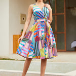 A woman modeling a sleeveless, knee-length wrap-style dress with a colorful geometric stripe pattern in pink, blue, yellow, green, orange, and purple, paired with red high heels, standing outdoors in front of a building.